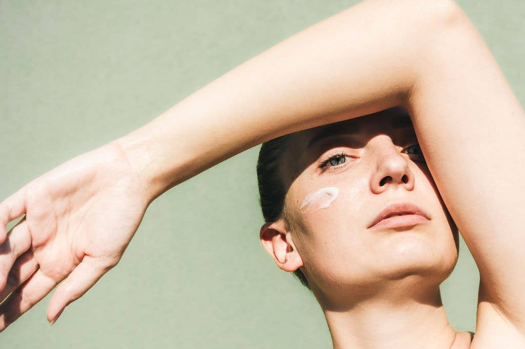 A young woman with a smear of cream on her cheek looks directly at the camera while shielding her eyes from the sun with her arm. This beautiful and authentic close-up captures a moment of self-care and confidence, highlighting a natural skincare routine.