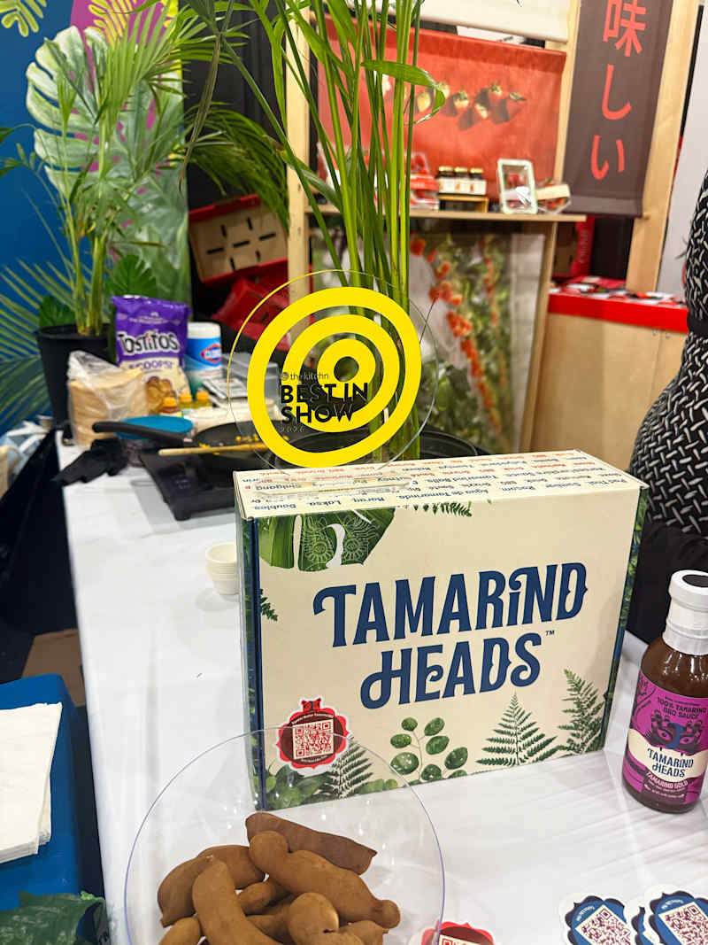 Tamarind Heads product box on a table with tamarind pods, a bottle of sauce, and tropical plants in the background.