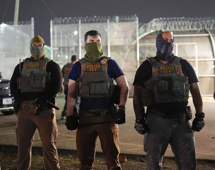 Three ICE officers in tactical gear stand in front of a fenced area at night. They wear masks and protective vests with "POLICE ICE" labels