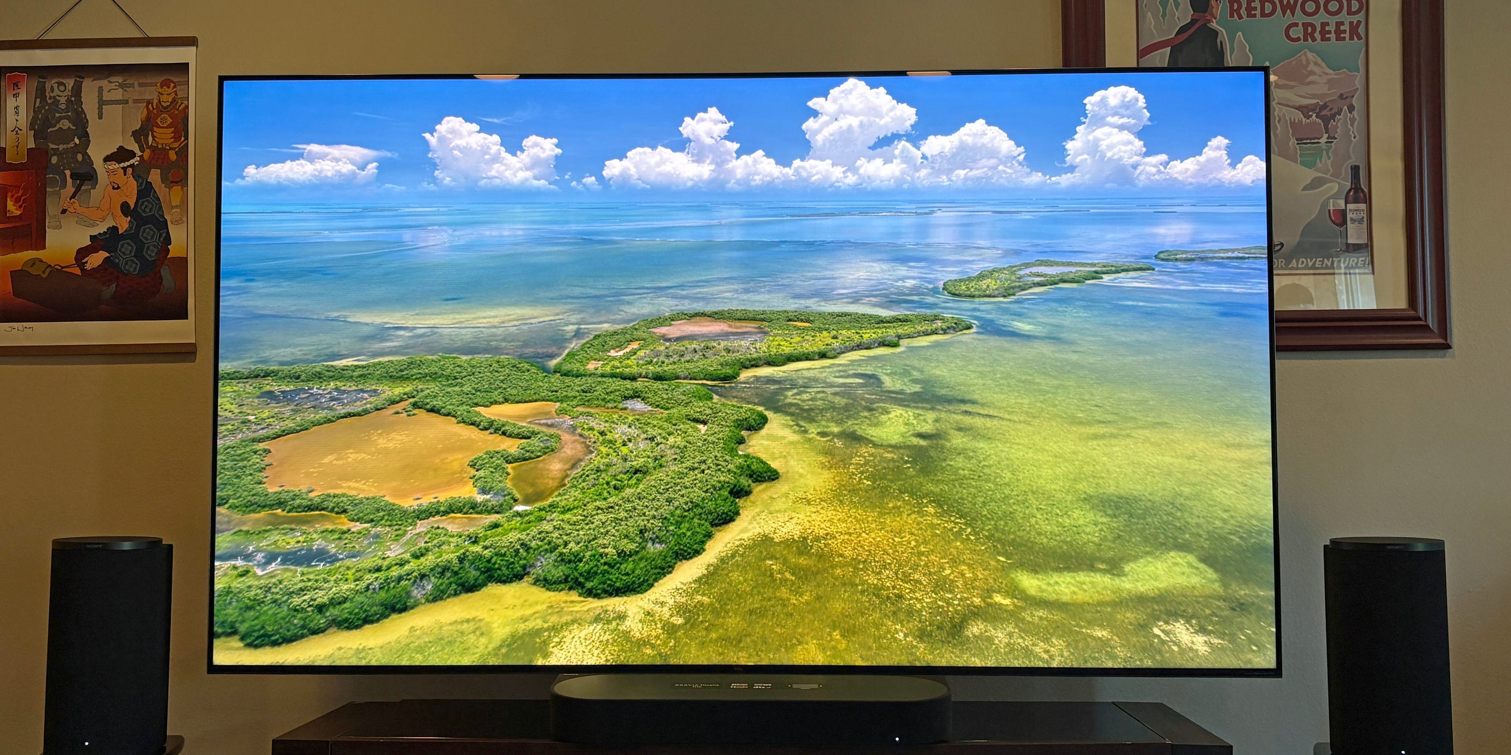 A TCL QM7L TV displays a vivid aerial coastal landscape in our reviewer's home entertainment setup with speakers and framed wall art.