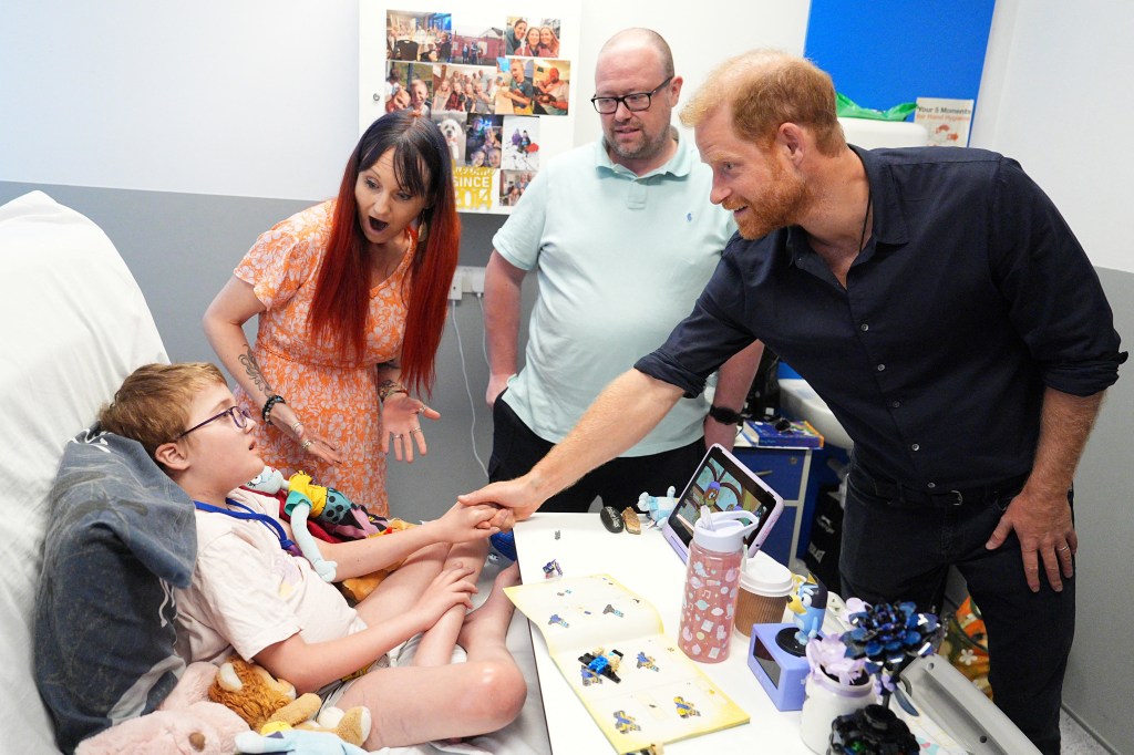 Prince Harry, in a navy blue shirt and black jeans, holds hands with patient Alec Hill, in a cream T-shirt, as his parents Kelly and Stuart Hill look on.
