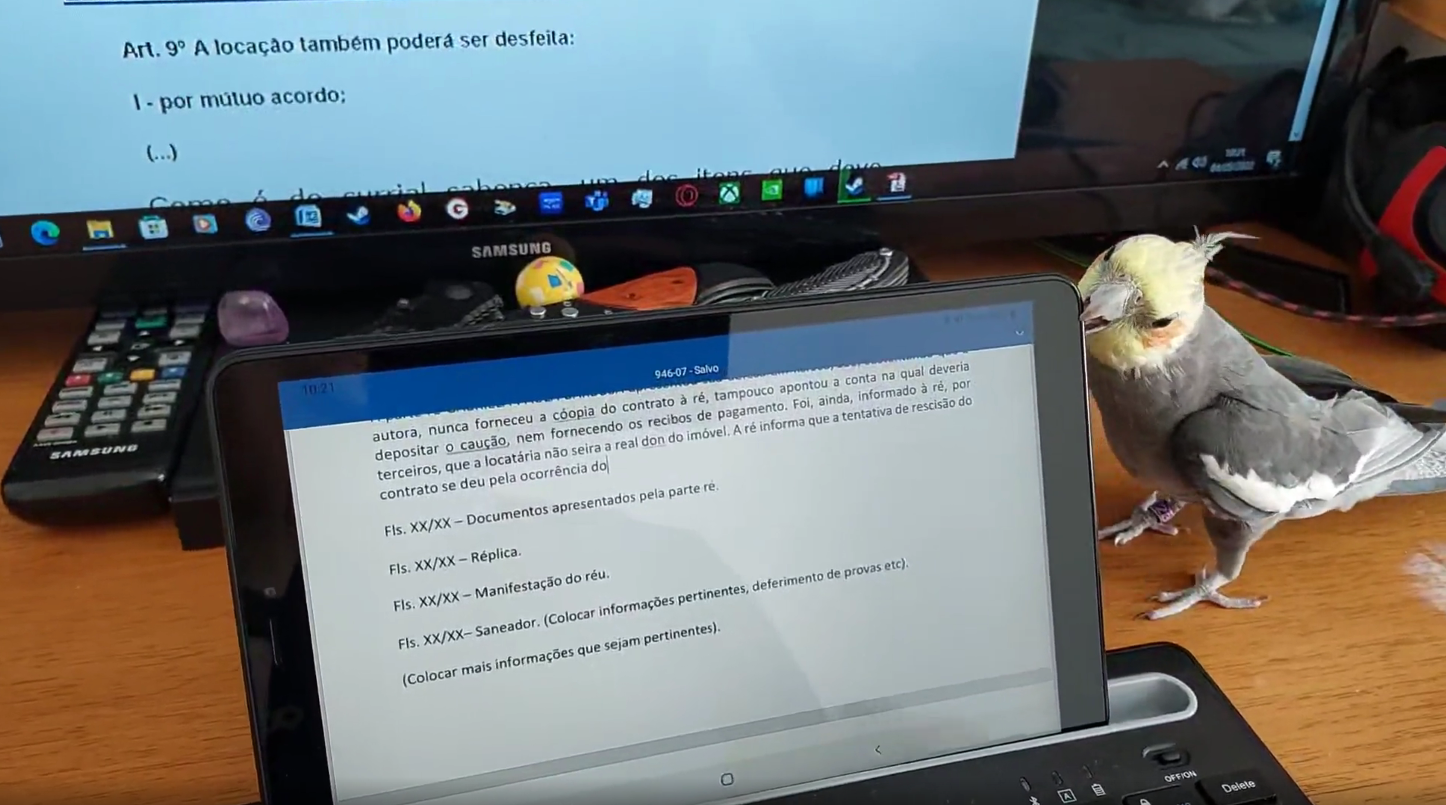 A cockatiel sits next to a tablet displaying a document with a monitor and remote in the background