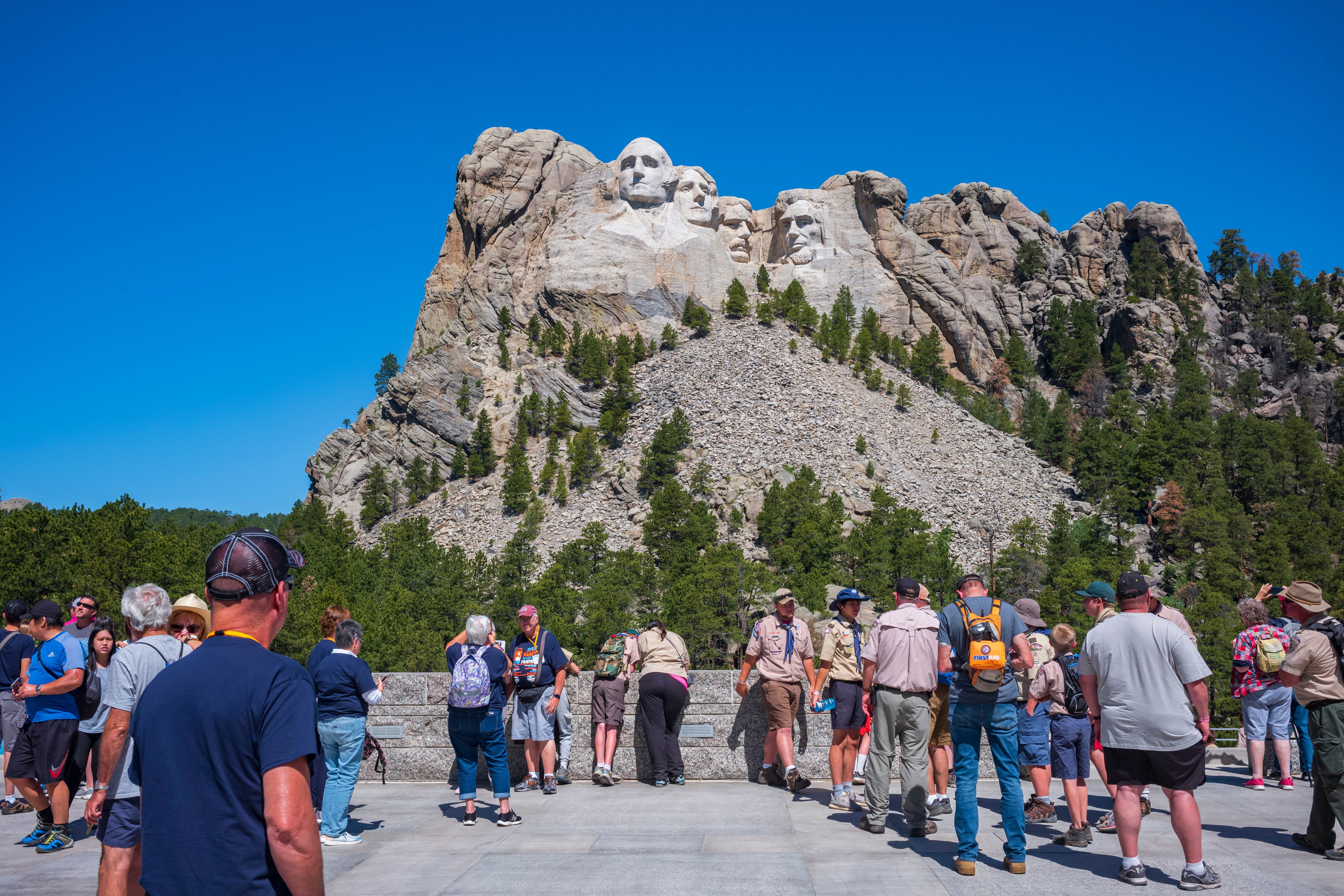 Tourists gather at Mount Rushmore, viewing the carved presidential faces on a sunny day