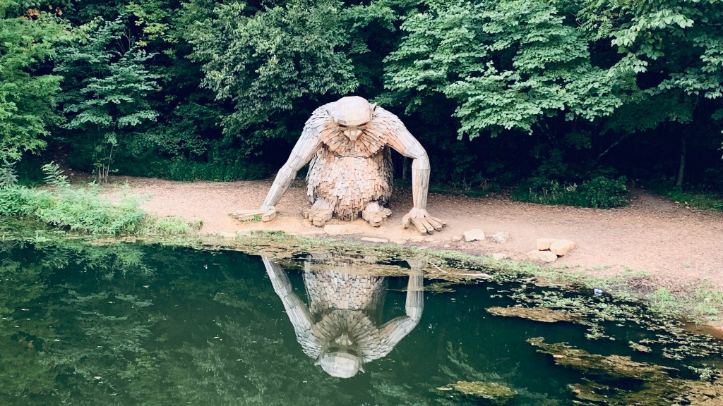 A smaller troll stands at the water's edge looking at its reflection in the pond.