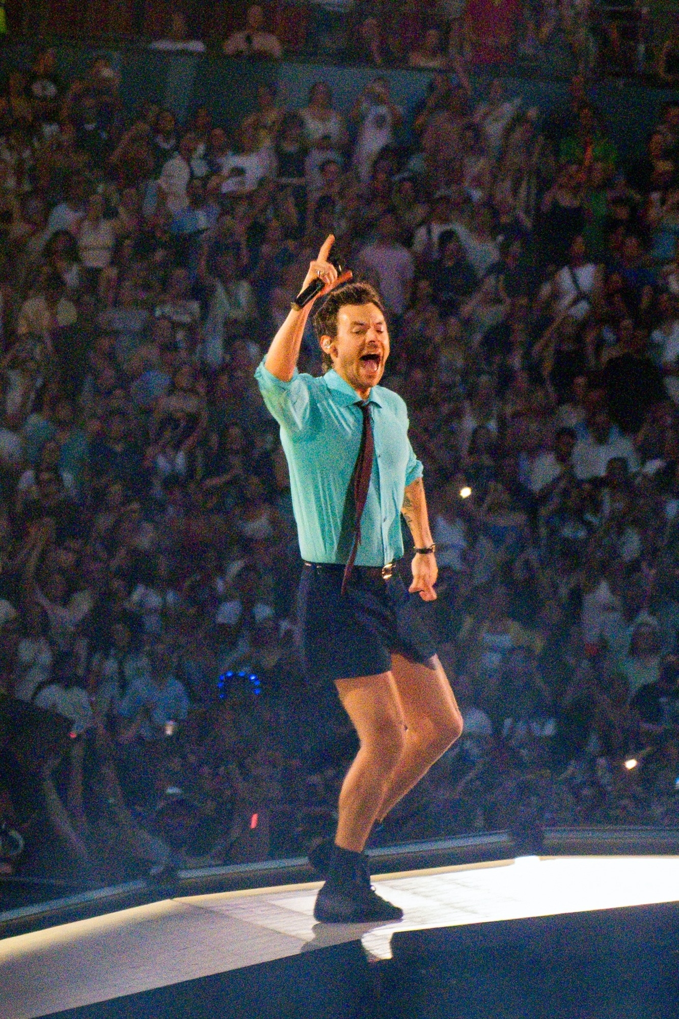 Harry Styles performing at Wembley Stadium, wearing a light blue shirt, dark shorts, and a tie, holding a microphone.