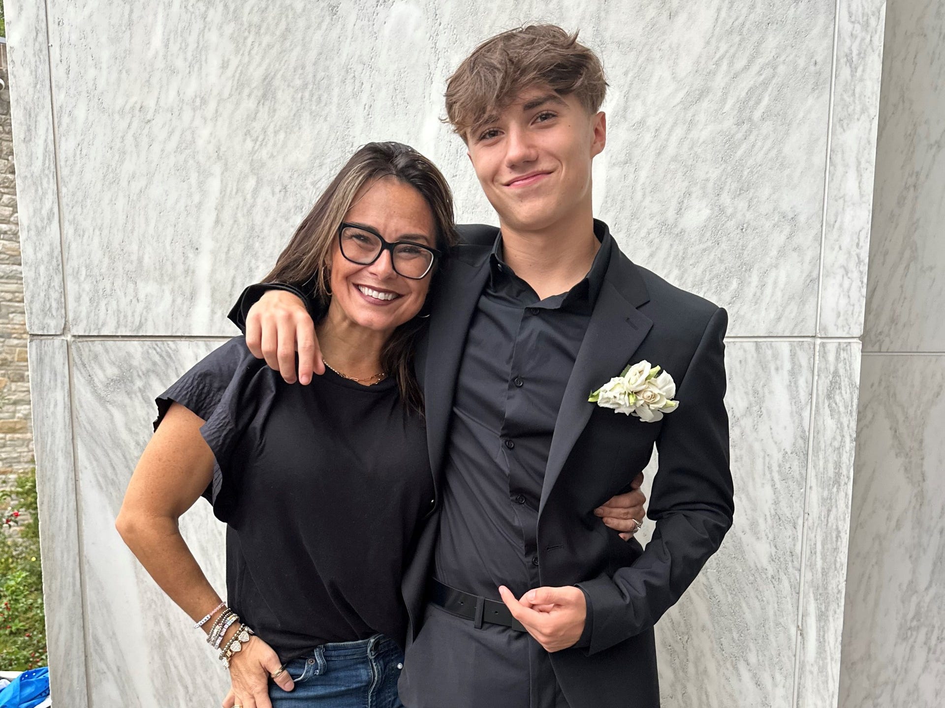Two people pose together in front of a marble wall, one wearing a black suit with a white boutonniere.