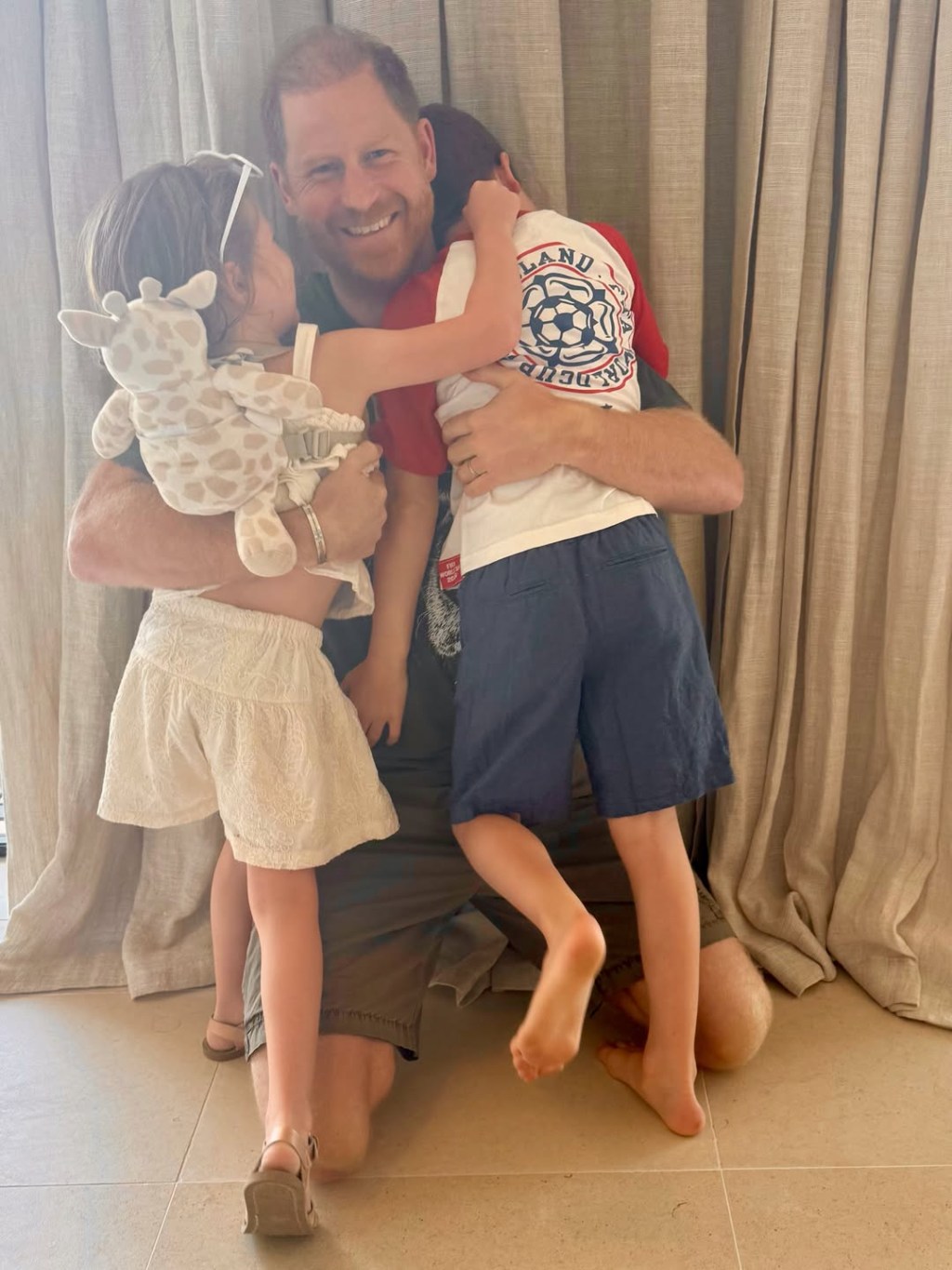 Prince Harry kneeling and embracing his children, Prince Archie, in a red and white England soccer shirt and blue shorts, and Princess Lilibet.in a cream top and cream socks.