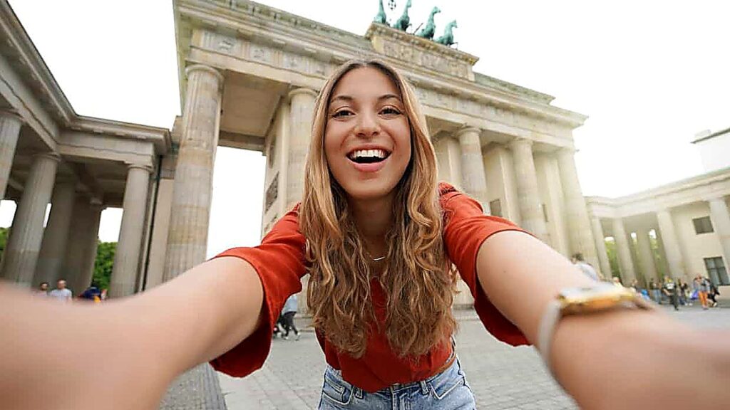 A woman with long hair smiles and takes a selfie in front of the Brandenburg Gate, a historic monument with columns and a quadriga statue in Berlin, Germany, capturing the moment during an exciting scavenger hunt.