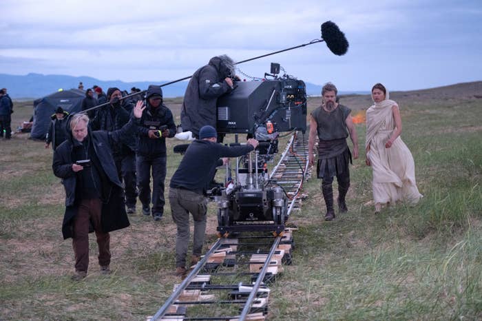 Film crew operates camera on tracks outdoors. Two actors in period costumes walk alongside, surrounded by vast, grassy landscape