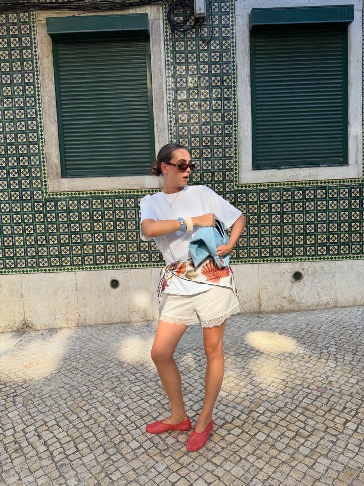 A person stands outdoors in front of a tiled wall and windows holding a lightweight bag and wearing sunglasses