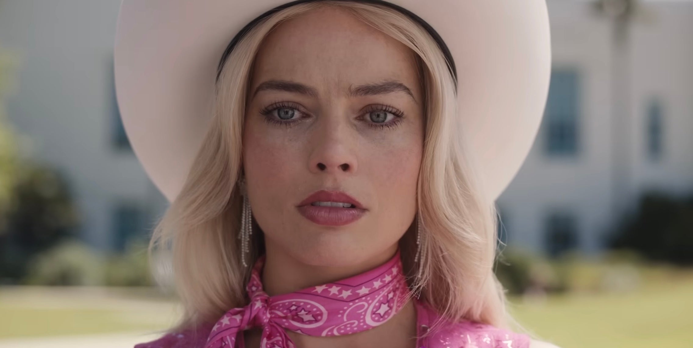 Margot Robbie as Barbie in the “Barbie” movie in a wide-brimmed hat, wearing a patterned scarf and earrings, looking forward with a neutral expression