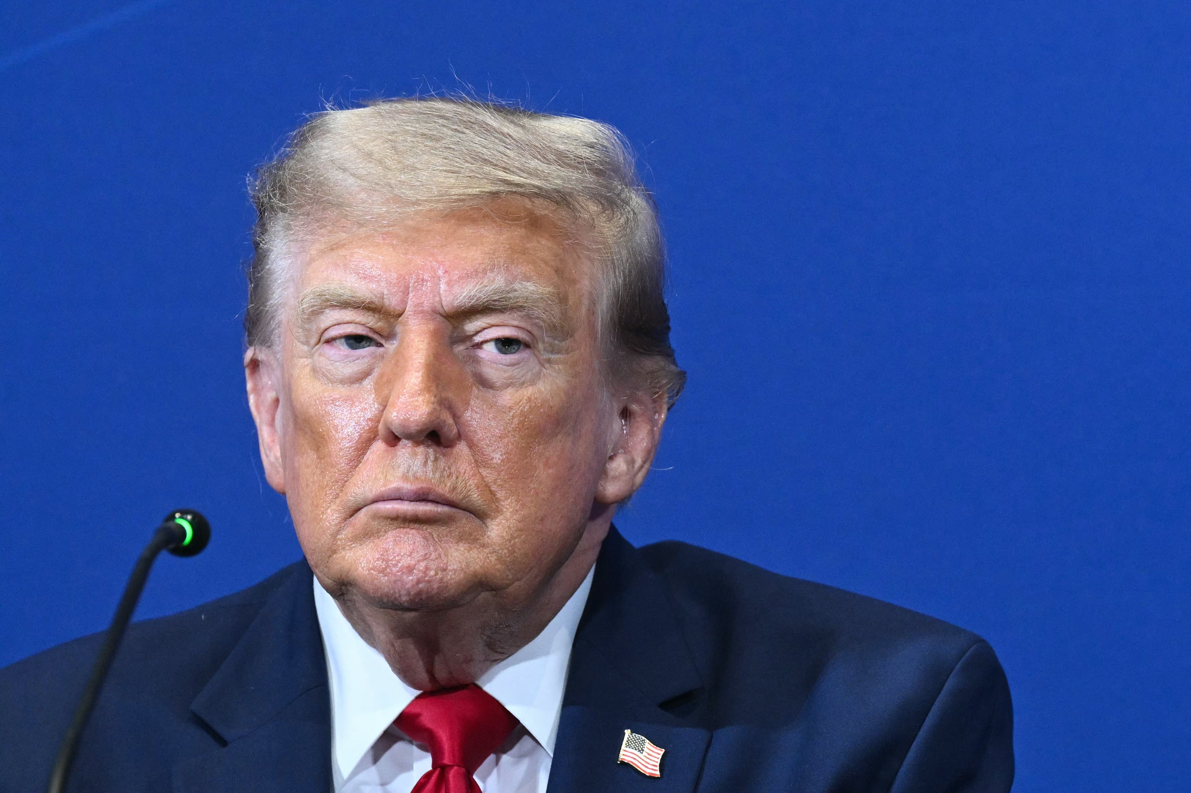 A person in a suit and tie sits at a blue background event, appearing thoughtful and attentive