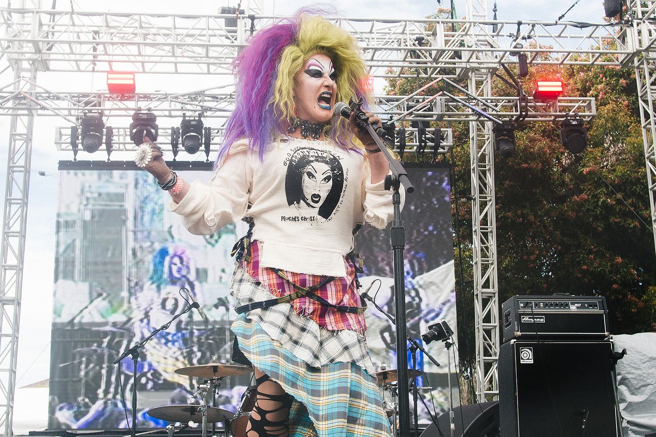 Peaches Christ performing on an outdoor stage with vibrant costume and makeup holding a microphone