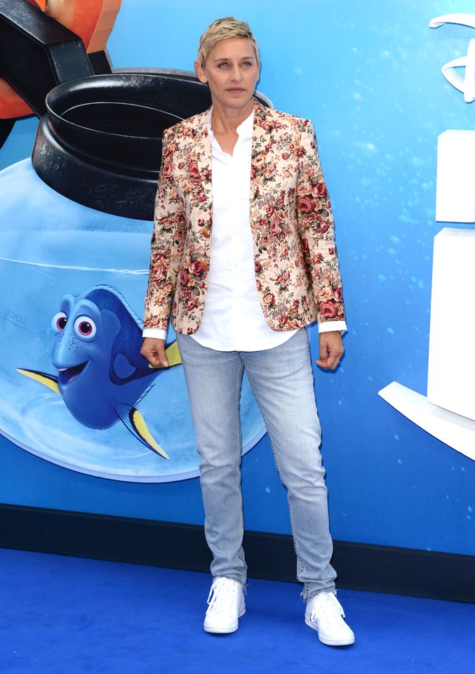 Ellen DeGeneres stands on a blue carpet in front of a backdrop with animated characters
