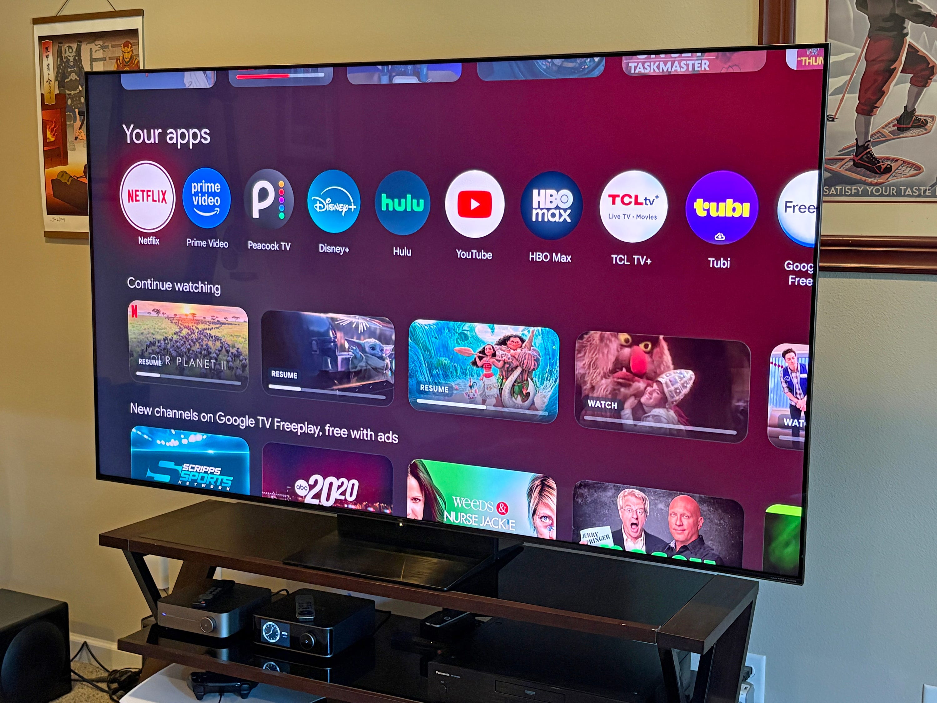 A TCL QM7L TV displays the Google TV home screen with streaming app icons and continue-watching rows.