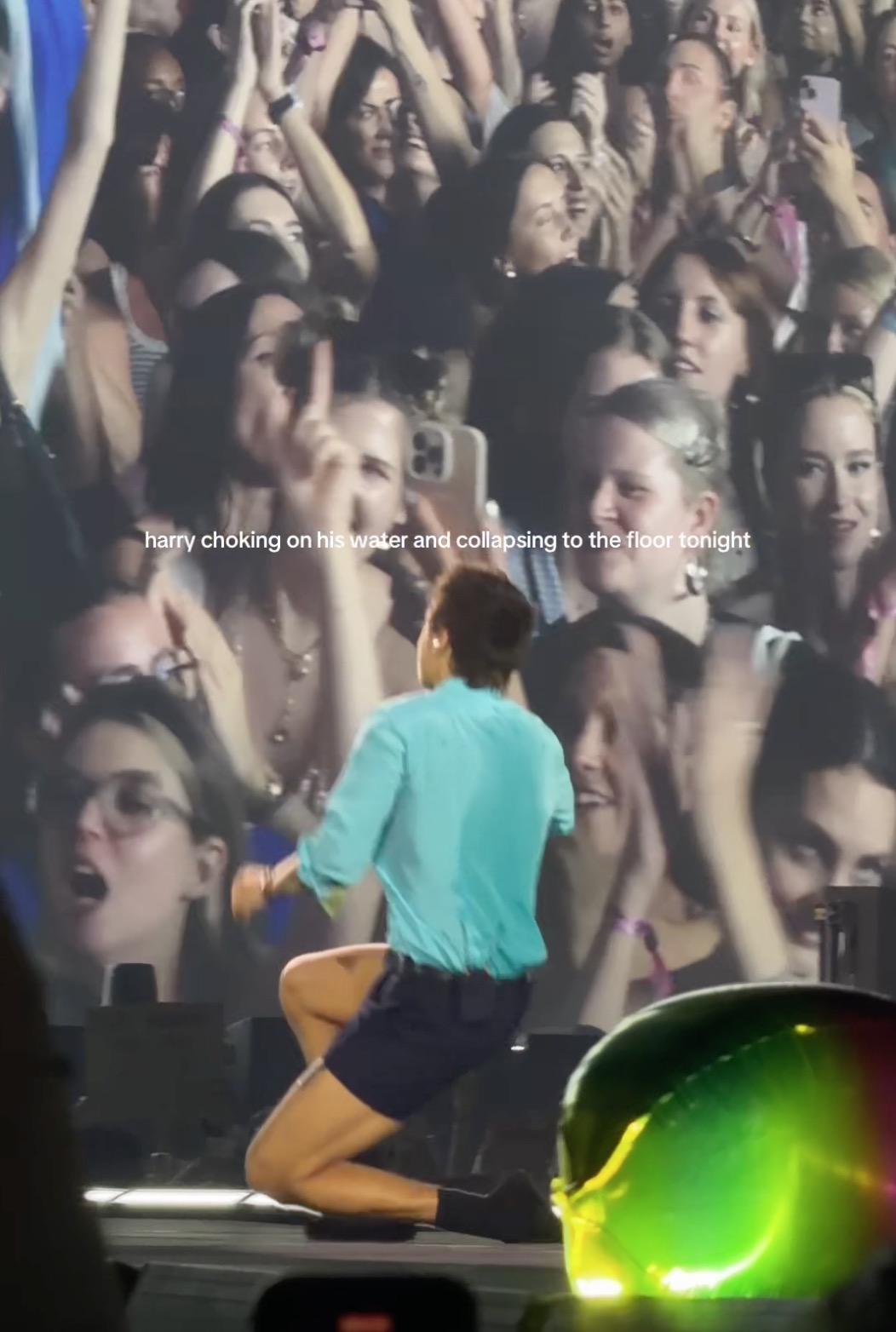 Harry Styles kneeling on stage with a crowd visible behind him.