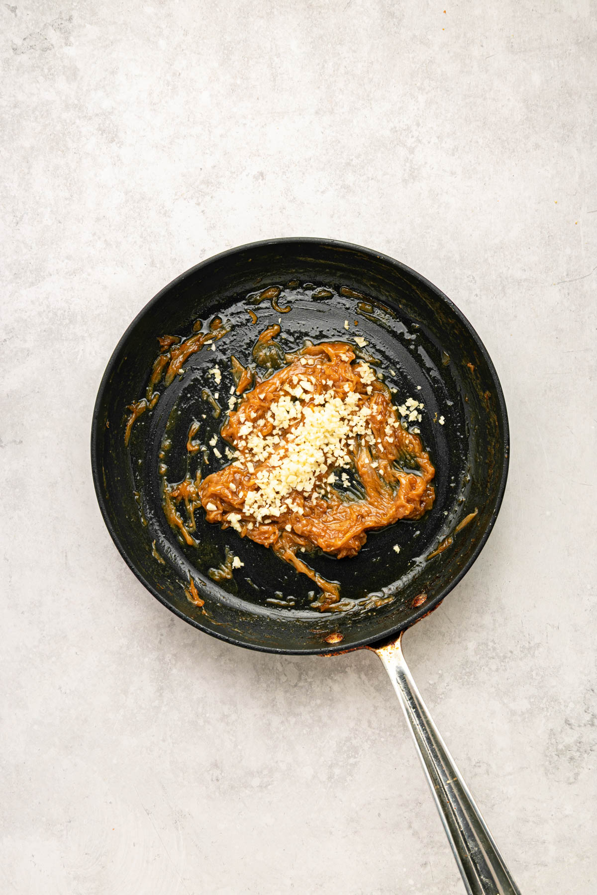 A black frying pan with caramelized onions and minced garlic sits on a light grey surface.