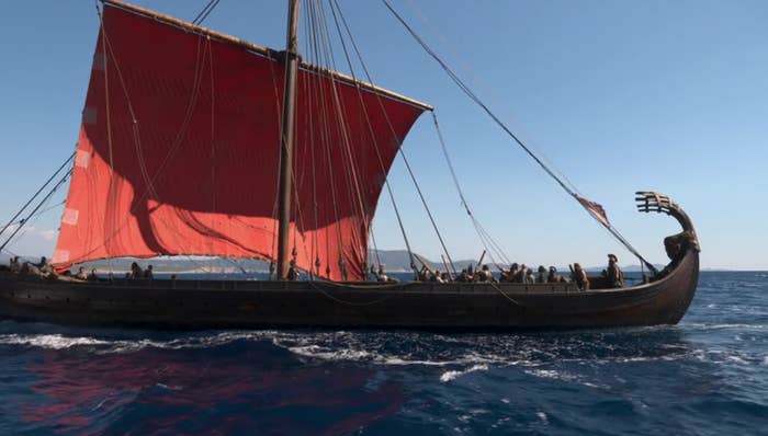 A Viking longship with a large sail and many people on board is sailing on the ocean