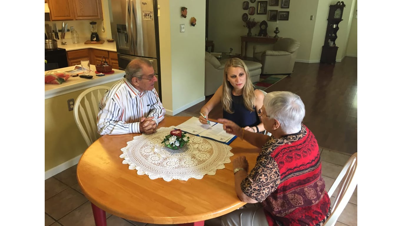 A blonde woman in a dark top sits at a round wooden table, holding a pen and looking at documents. She is engaged in a conversation with an elderly man wearing a striped shirt and an elderly woman in a red patterned top who is pointing with her hand. The table is covered with a white lace doily and features a small floral centerpiece. In the background, a kitchen with wooden cabinets and a living room area are visible.