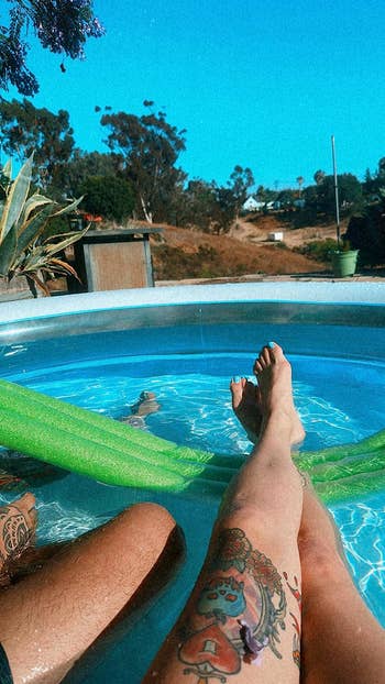 Person relaxing with legs in an inflatable pool, surrounded by greenery and plants. A tattoo is visible on one leg
