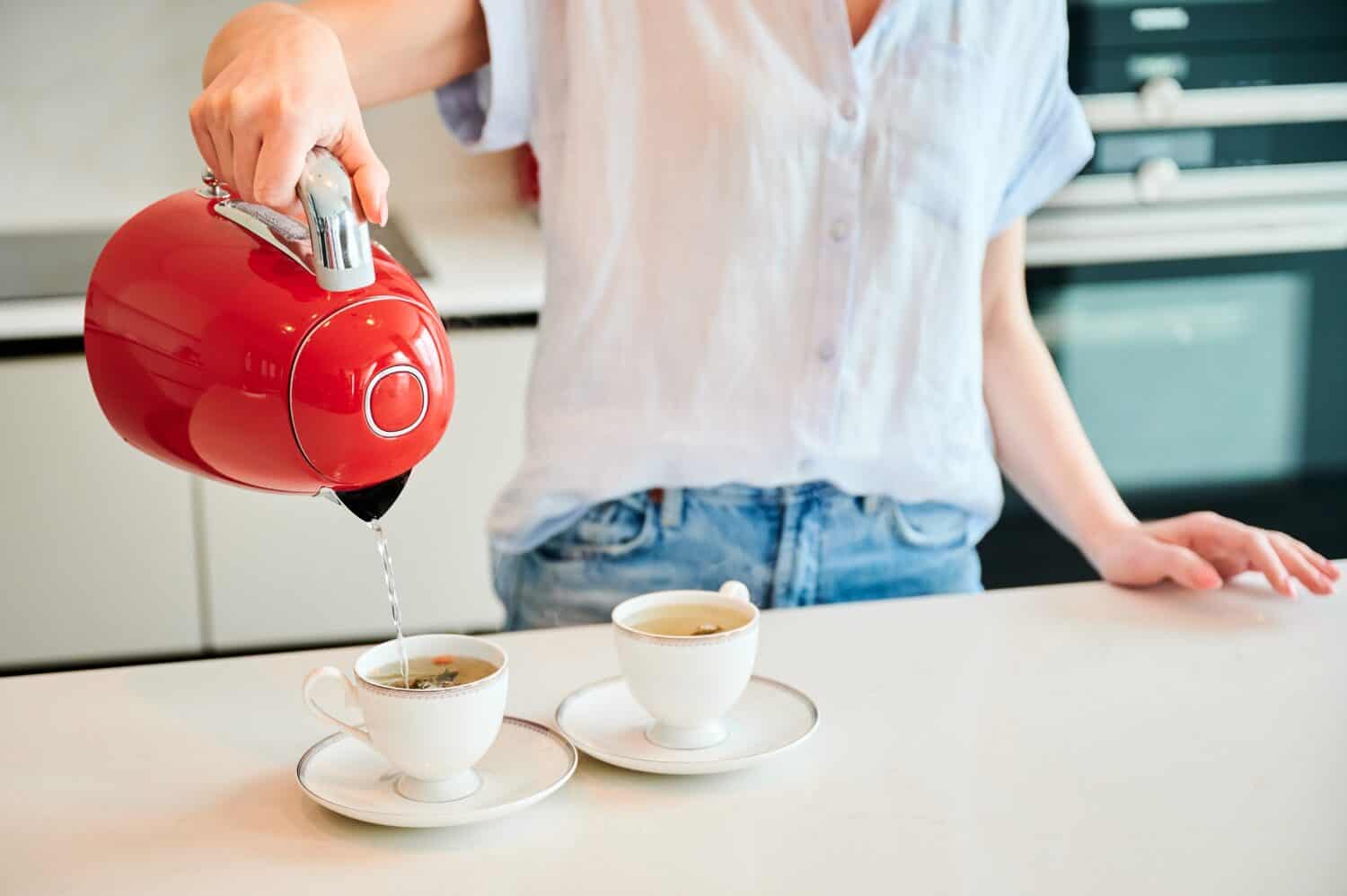 Cropped picture of woman pouring water into cup. Young female making tea for two. Woman holding red kettle over two cups.