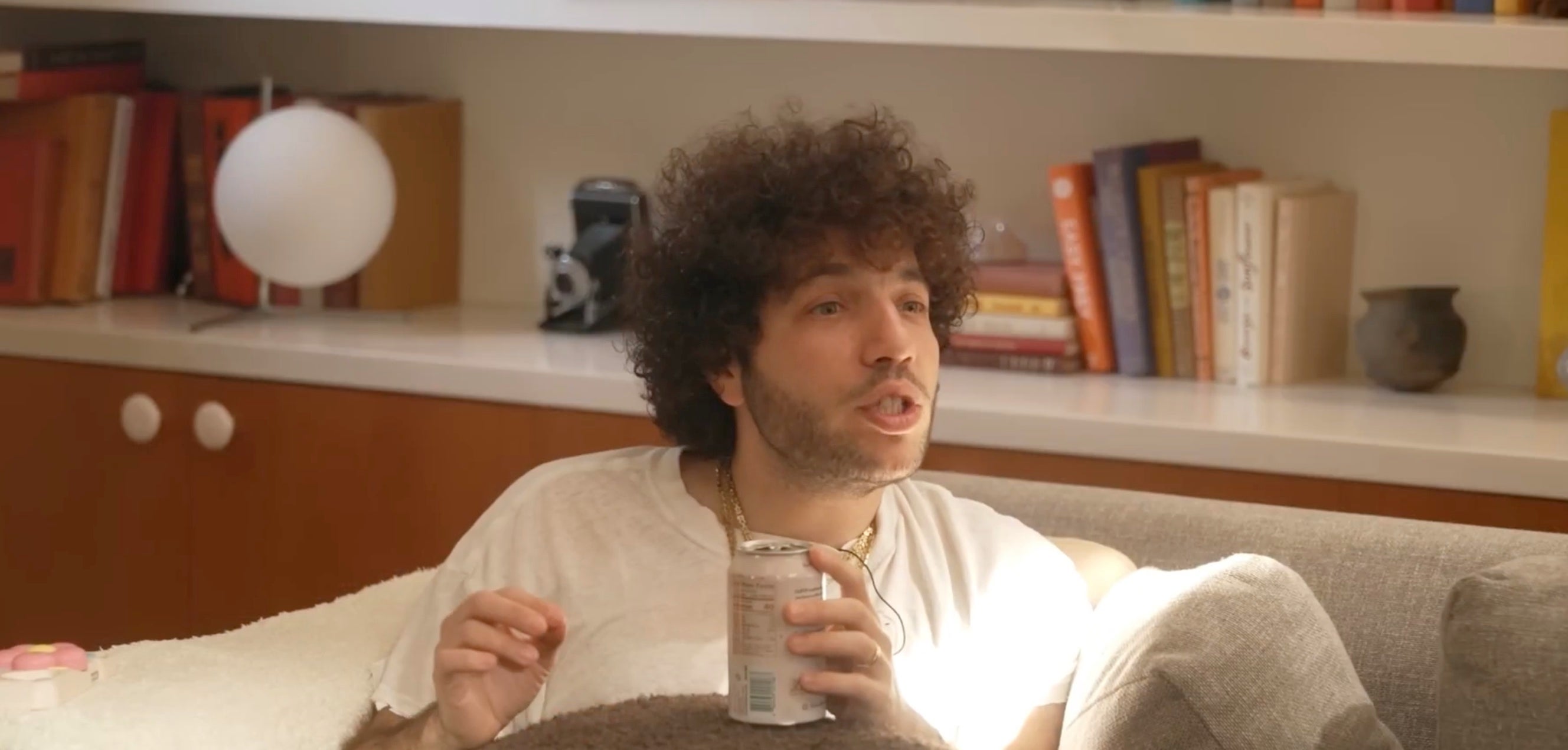 Person with curly hair sits on a couch holding a can, speaking animatedly, with books and a lamp in the background