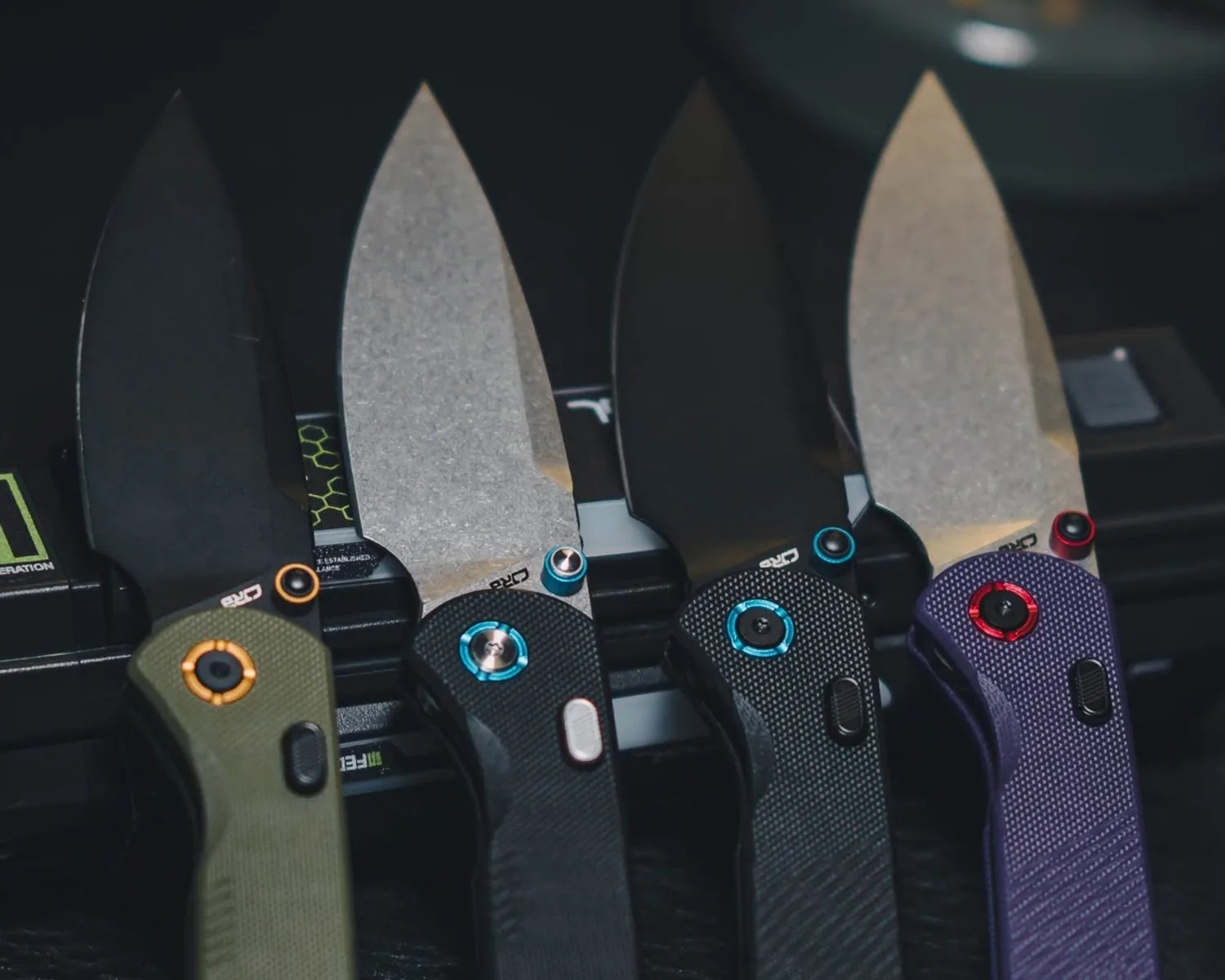 Four folding knives with textured handles in green, black, black, and purple, each with partially serrated blades and colored accents on the pivot screws.