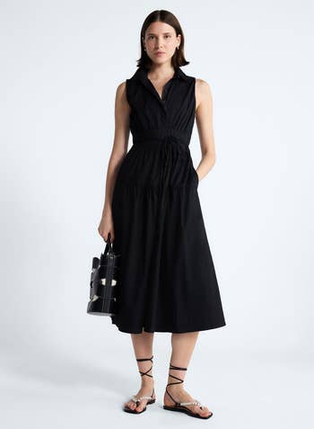 Person in a sleeveless black dress with tied waist, holding a stylish handbag, wearing strappy sandals, posed for a shopping catalog