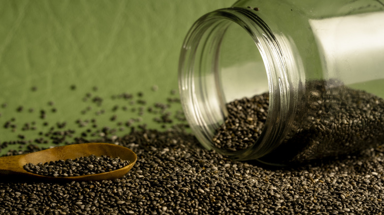 A jar of of chia seeds next to a pile of its contents spilled on a green surface.
