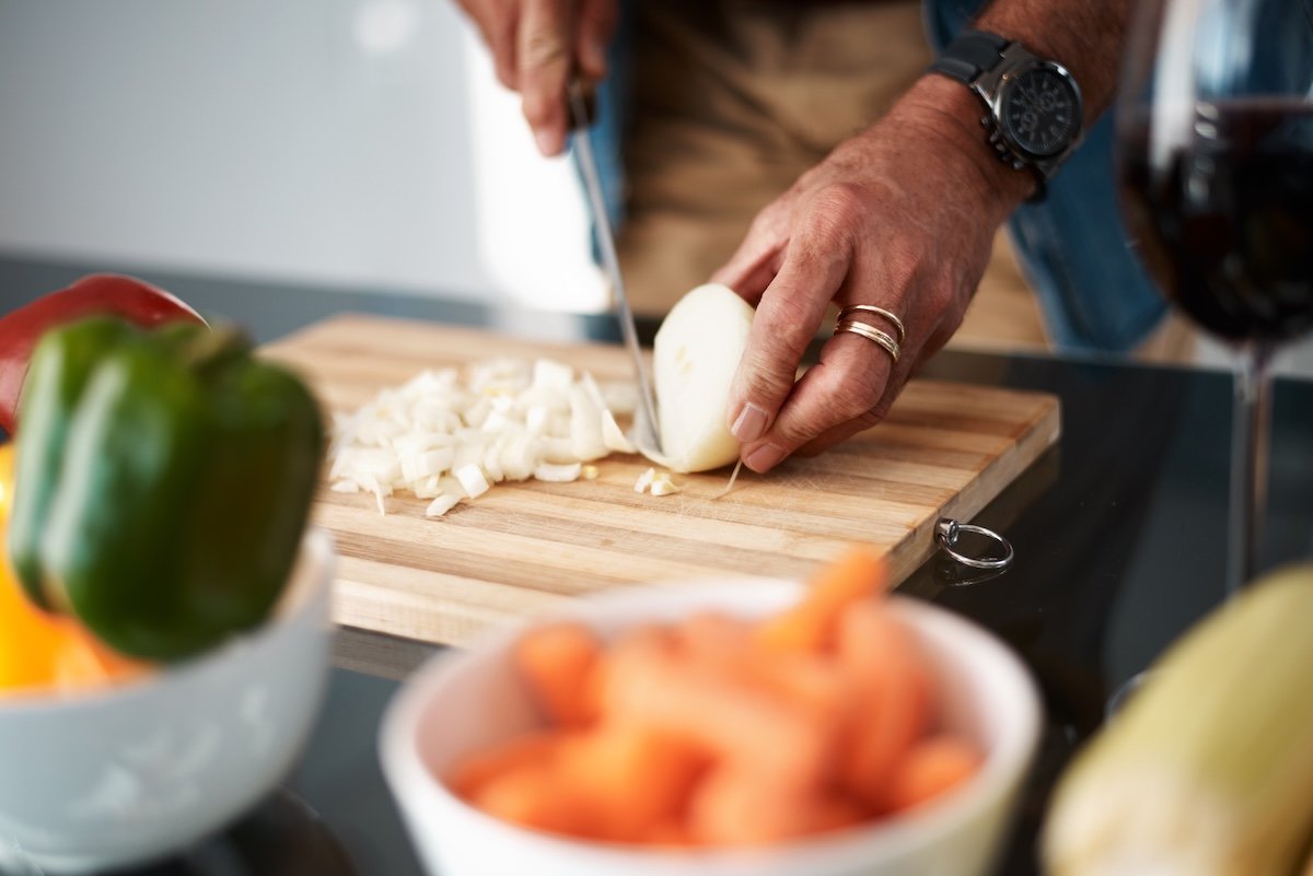 A man wearing rings and a watch chops a yellow onion on a wooden cutting board.