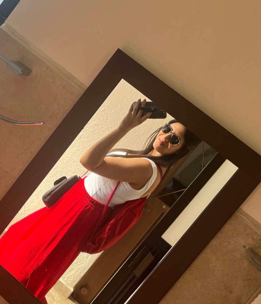 A traveler wearing a white off-the-shoulder top, flowing red skirt, sunglasses, and a matching red shoulder bag takes a mirror selfie in warm natural light. The casual travel outfit is reflected in a full-length mirror inside a modern room.