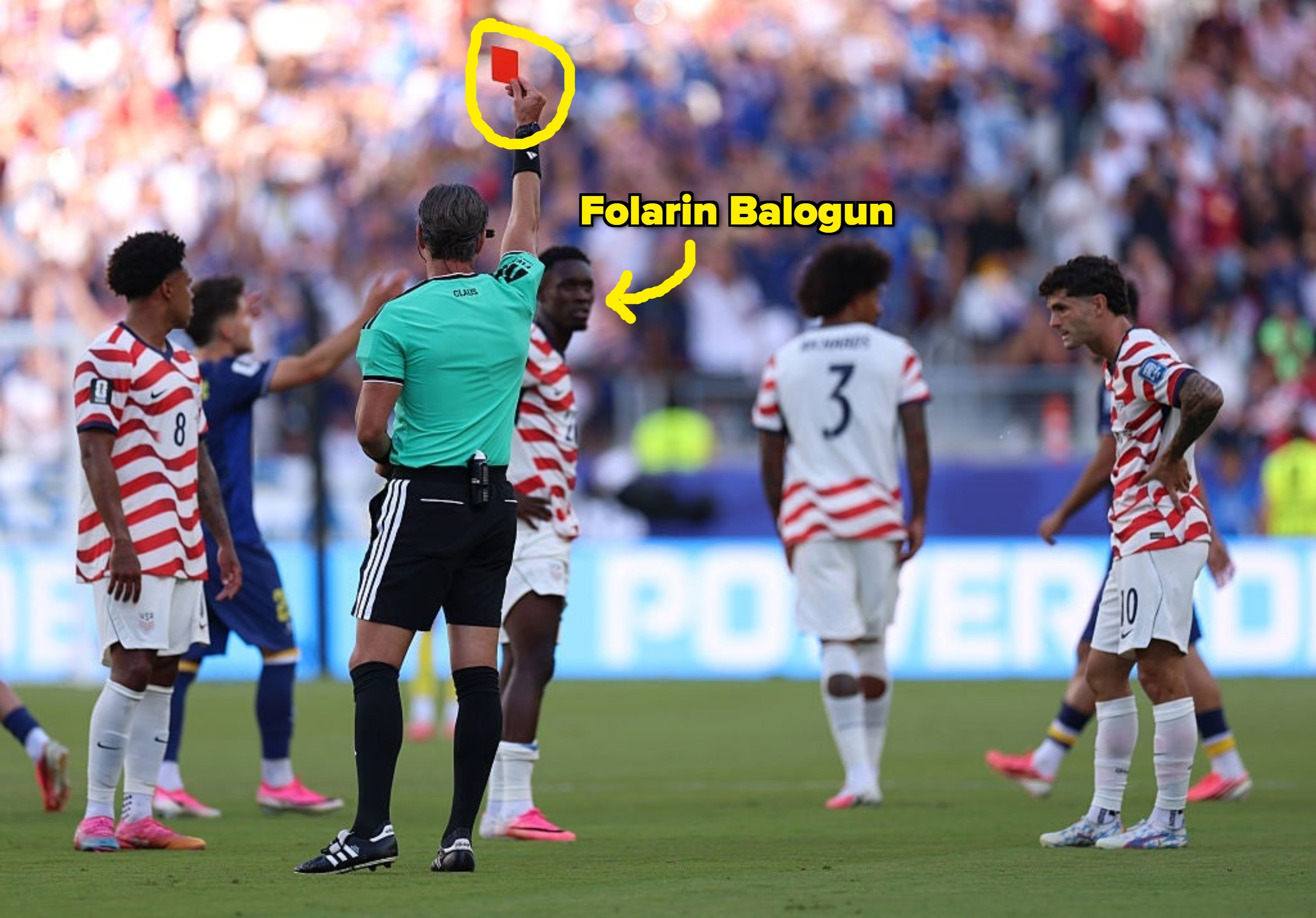 A soccer referee holds up a red card as American players react on the field during a match