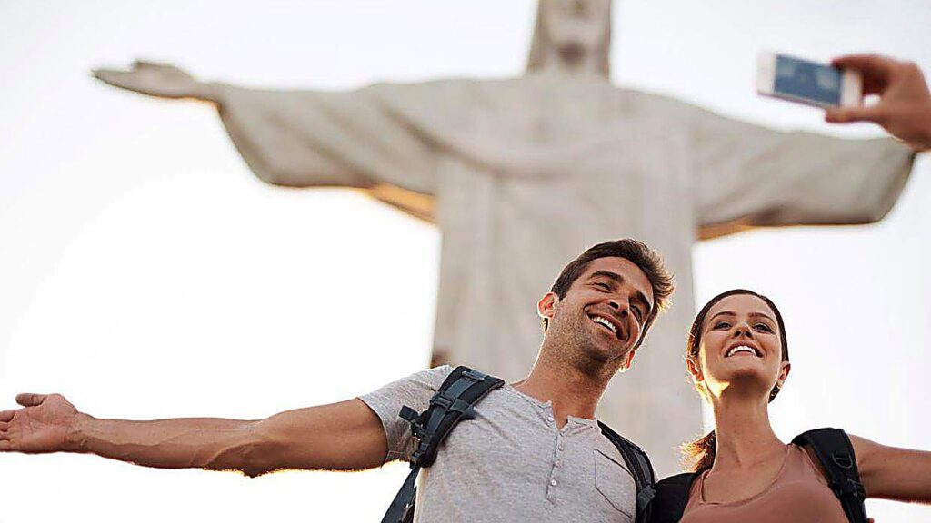 Two people with backpacks smile and pose for a selfie in front of the Christ the Redeemer statue in Rio de Janeiro, capturing a moment from their exciting scavenger hunt adventure.