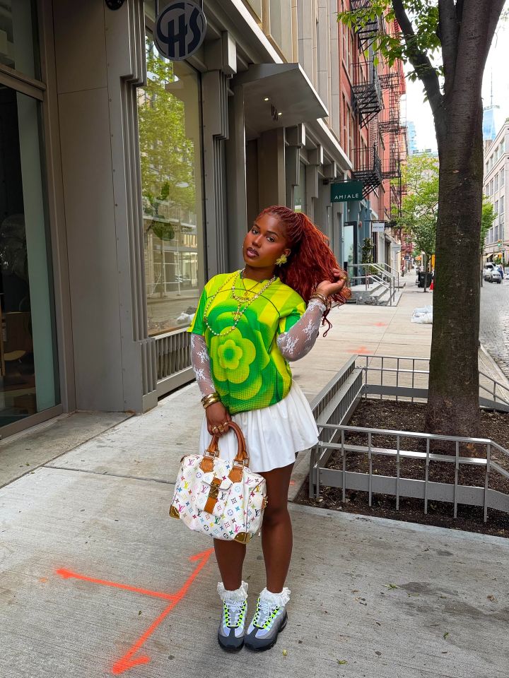 Person standing on a city sidewalk wearing a floral shirt white skirt and sneakers carrying a bag