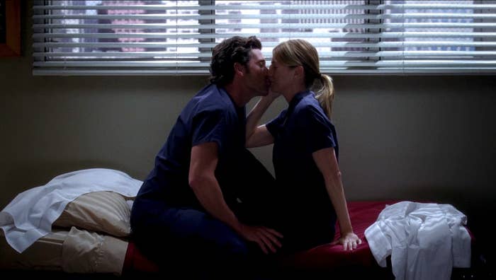 Two people in medical scrubs share a kiss while sitting on a bed in a dimly lit room with blinds partially open