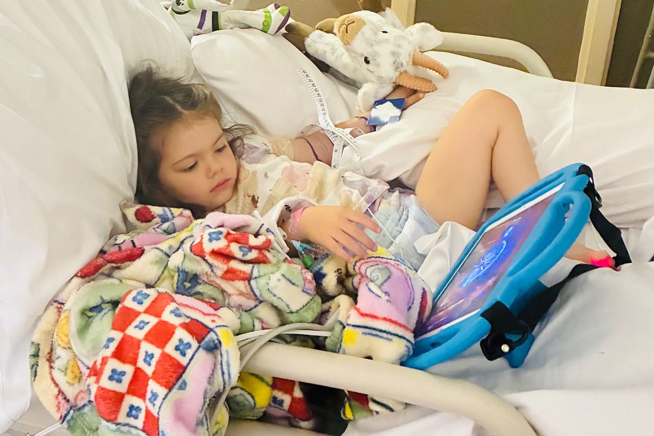 A young girl lying in a hospital bed using a tablet surrounded by toys and a colorful blanket