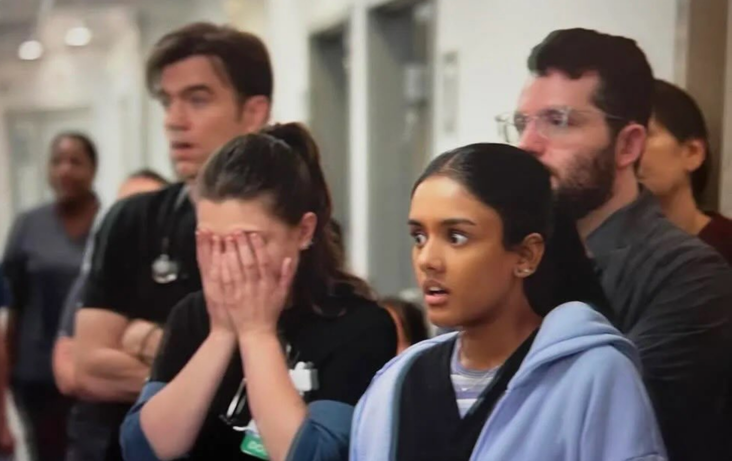 People standing indoors, looking surprised or shocked. One person covers their face with their hands