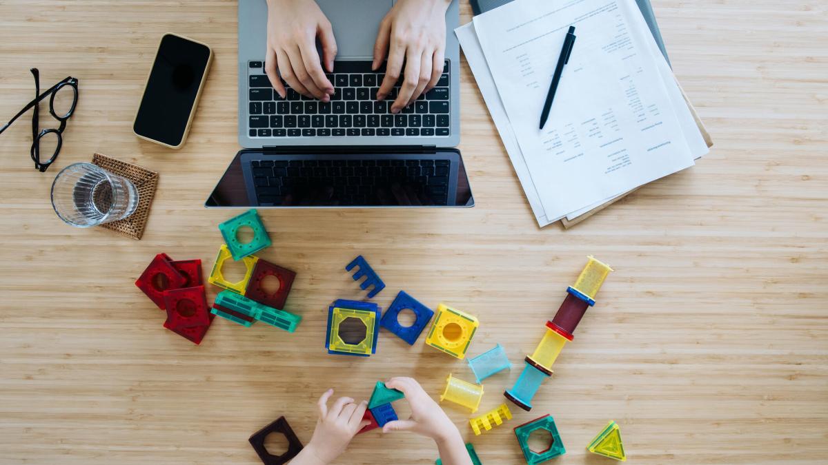 An image showing a woman working on a laptop while a child plays with blocks in front of her