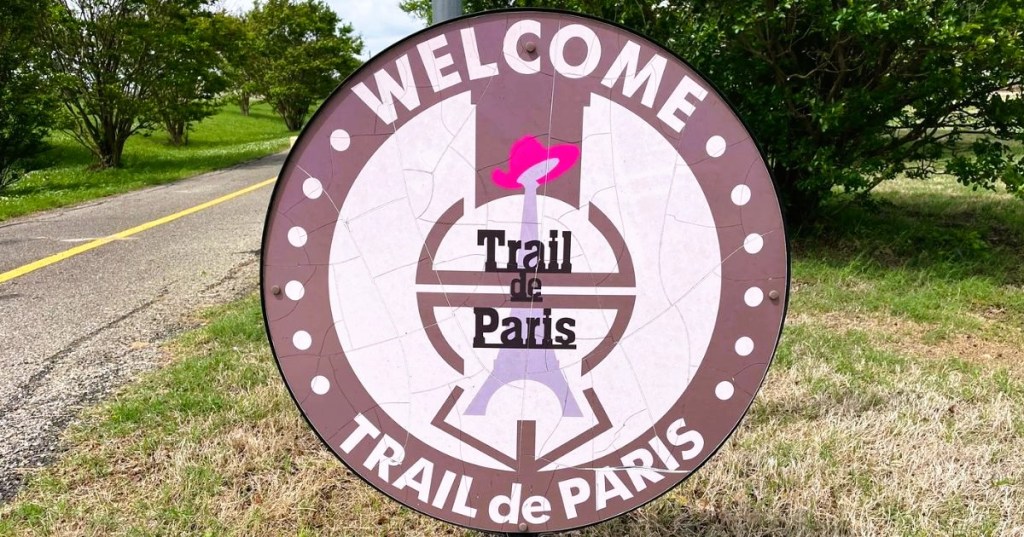 Trailhead welcome sign for Trail de Paris, a rail-to-trail paved path near Dallas, Texas, surrounded by trees.