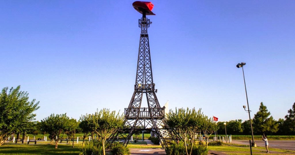 A traveler taking pics of a replica of the Eiffel Tower in Paris, Texas, with a large red cowboy hat on top, surrounded by greenery.