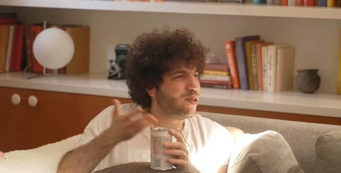 Person with curly hair sitting on a couch, holding a canned drink, gesturing as if in conversation, books and decor in the background