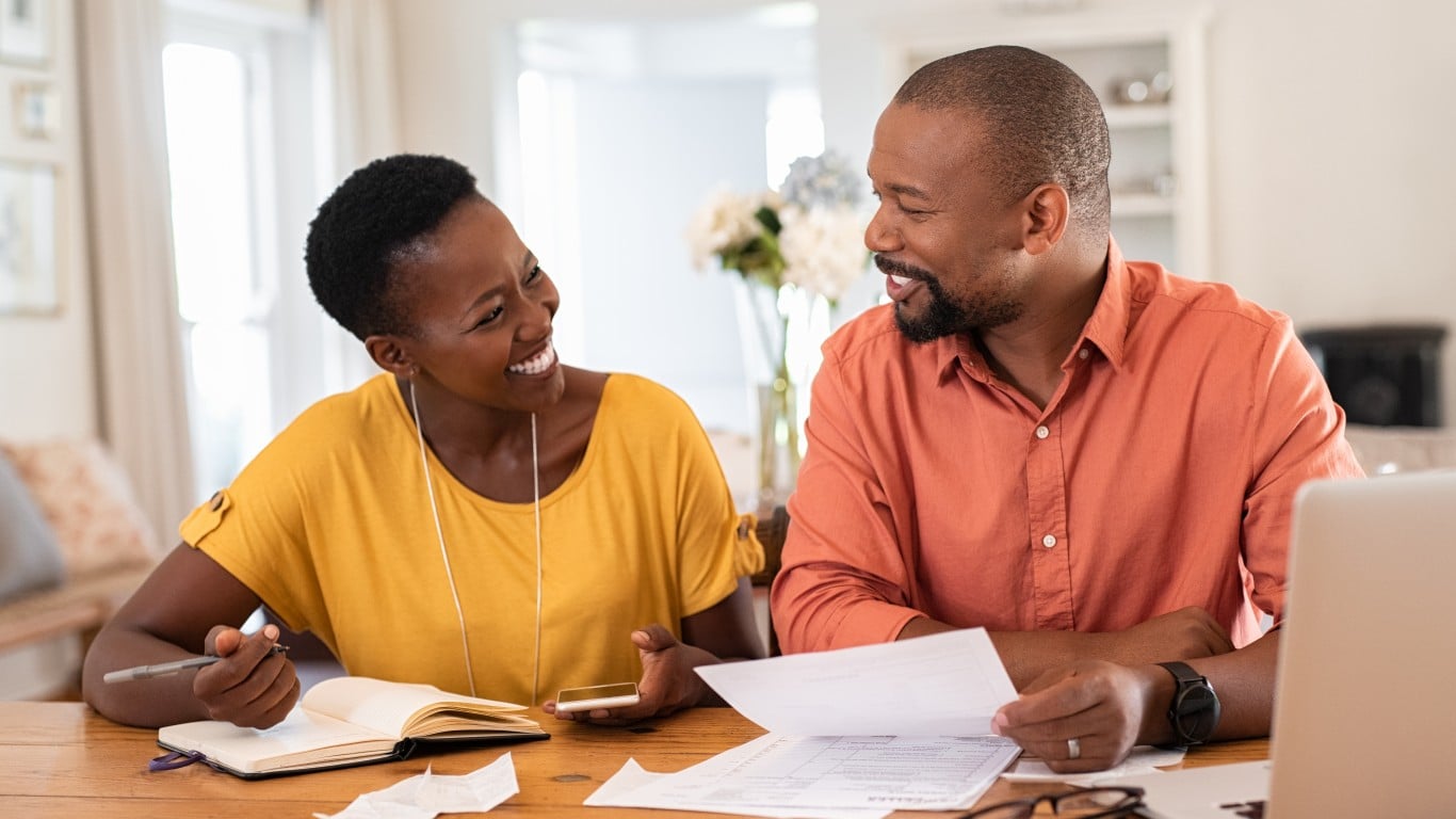 A Black man and woman sit at a wooden table, smiling and looking at each other. The woman in a yellow shirt holds a pen over an open notebook, while the man in an orange shirt holds financial papers. A laptop and eyeglasses are also visible on the table.