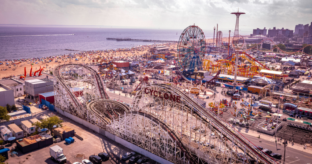 An overhead view of the theme parks at Coney Island, with roller coasters and ferris wheels not far from the beach.