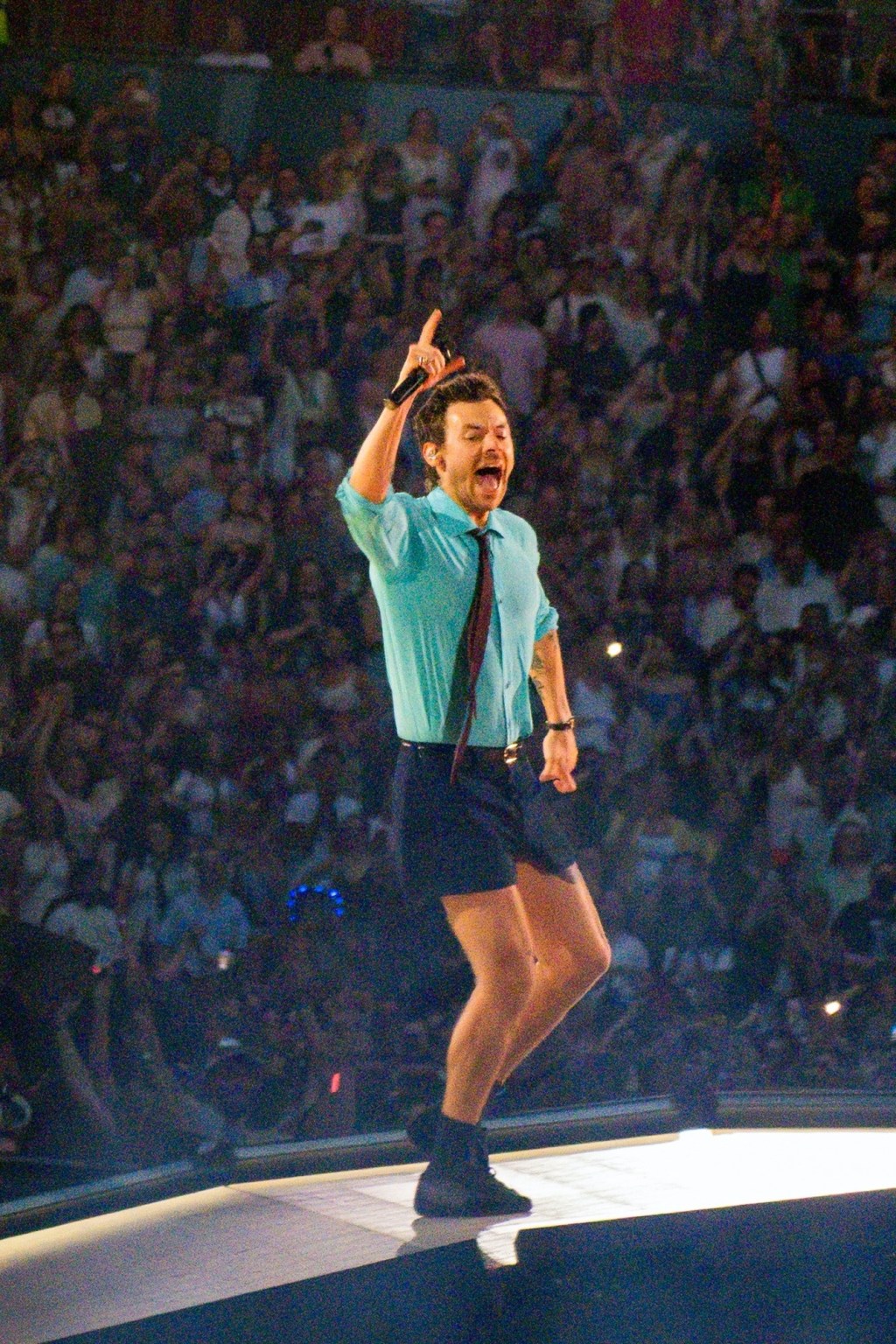 Harry Styles performing at Wembley Stadium, wearing a light blue shirt, dark shorts, and a tie, holding a microphone.