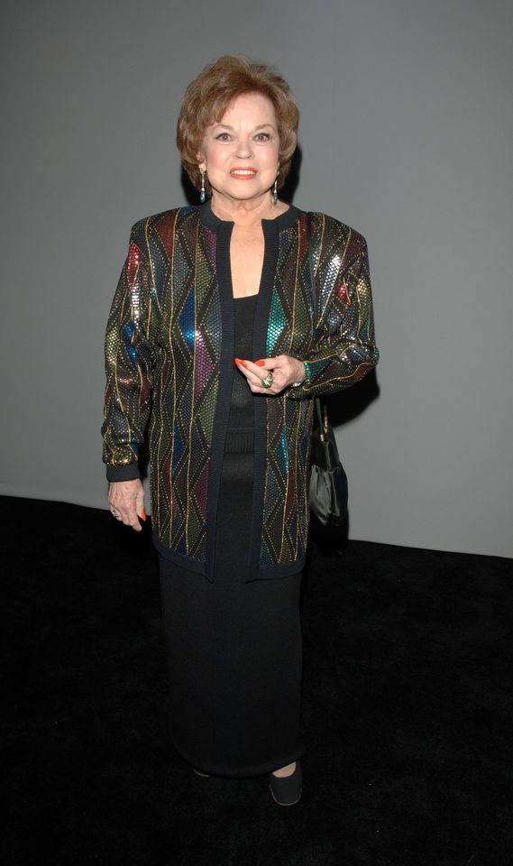 A person wearing a patterned jacket and standing on a black backdrop holding their hand partially extended