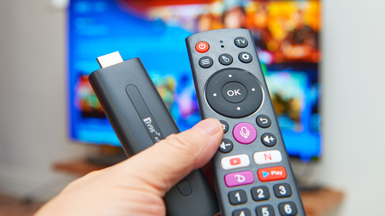 A hand holding a Fire TV Stick and remote with a blurred TV in the background.