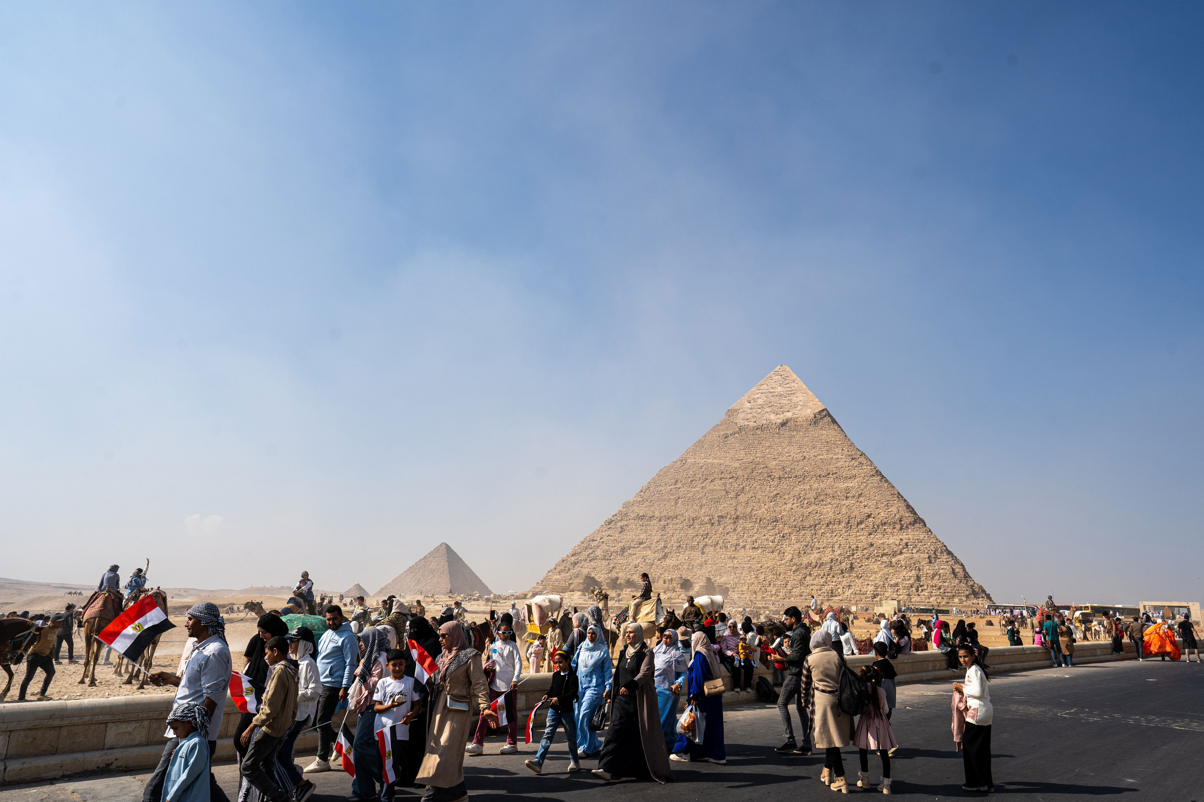 Tourists explore the area around the Pyramids of Giza, with camels and guides visible under a clear sky