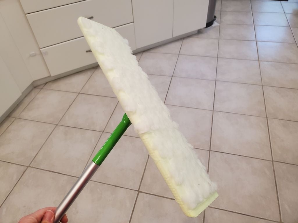 A hand holding a Swiffer-style mop with a fluffy cleaning pad in a tiled kitchen