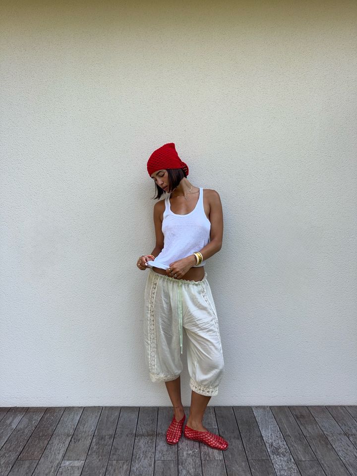 A woman wearing a red hat white tank top and loose pants standing by a wall on a wooden floor