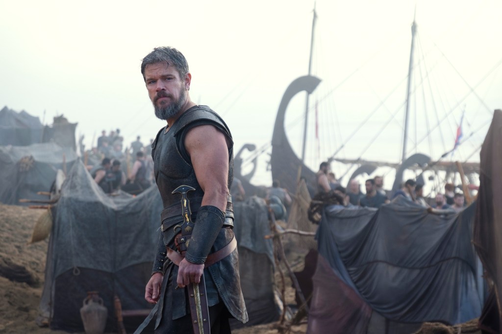 Matt Damon as Odysseus in armor holding a sword, with ships and a camp in the background.